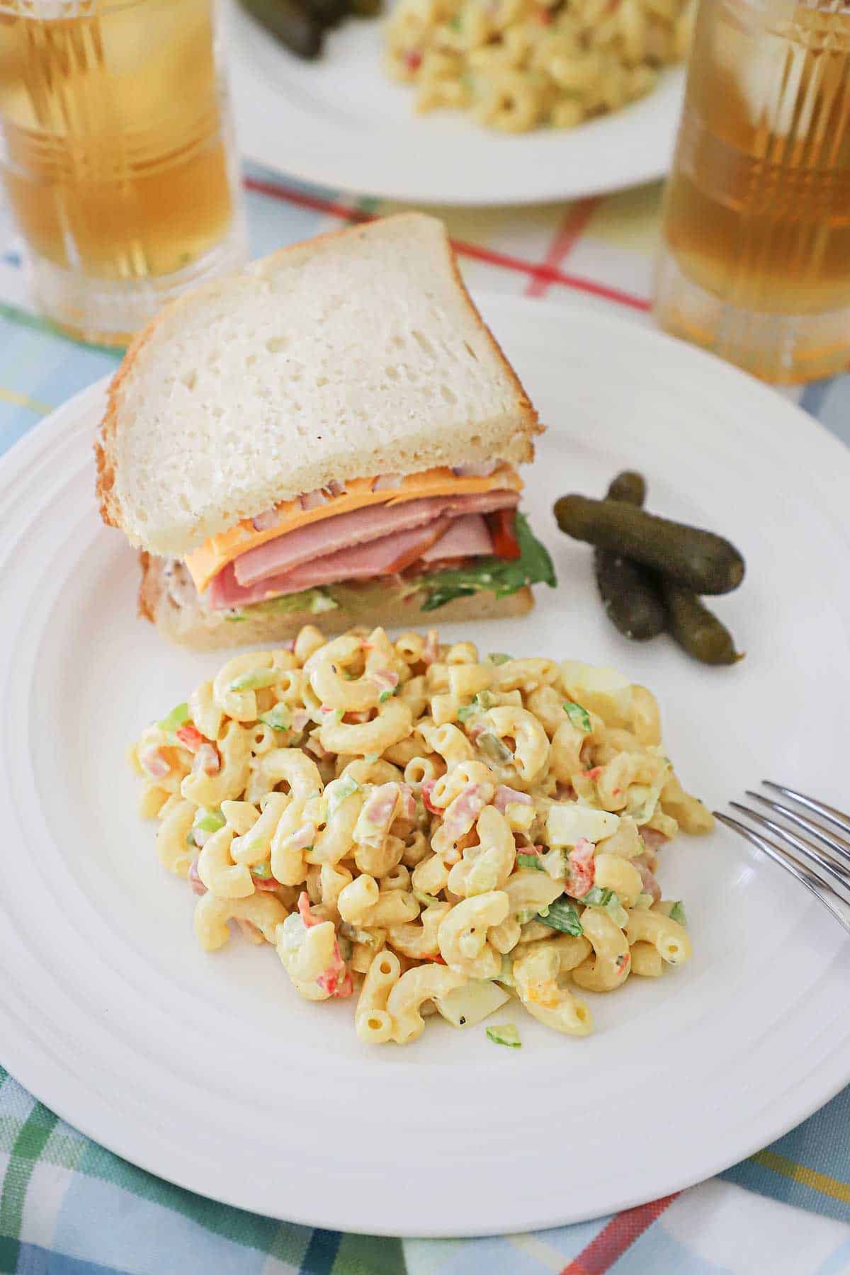 A white dinner plate filled with a serving of classic macaroni salad and half of a ham sandwich with three small pickles also sitting on the plate.