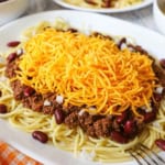 An oval white plate filled with Cincinnati chili served 5 ways including the chili over cooked spaghetti along with red kidney beans, chopped onion, and topped with a large mound of finely shredded cheddar cheese.