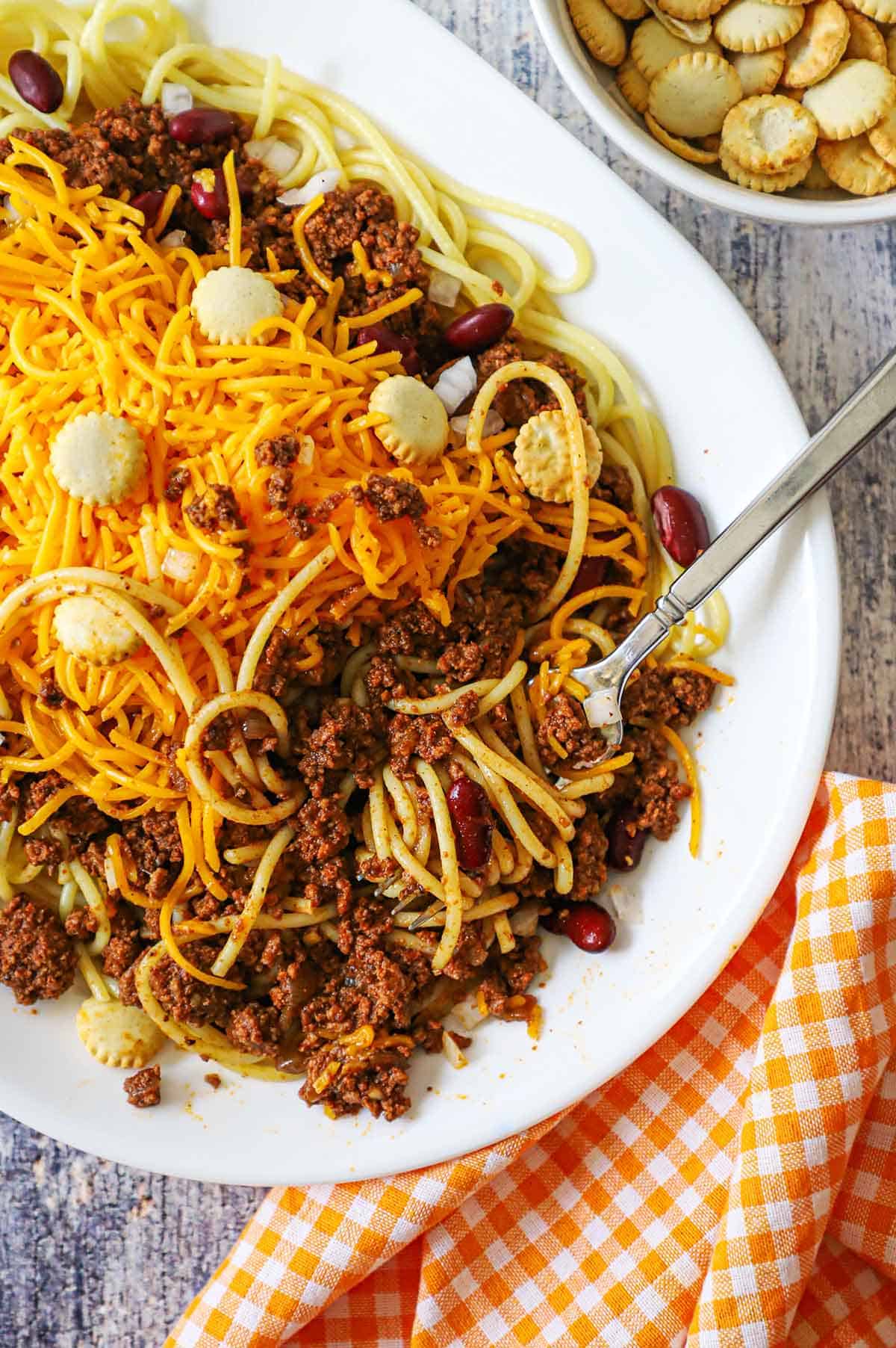A fork inserted into a large serving of Cincinnati chili on an oval white dinner plate filled with the chill over pasta and topped with a large mound of shredded cheddar cheese and oyster crackers.