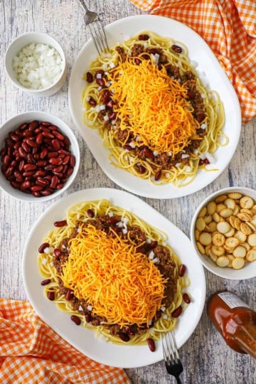 An overhead view of two large oval dinner plates filled with heaping servings of Cincinnati chili served over spaghetti, beans, onions, and topped with shredded cheddar cheese.
