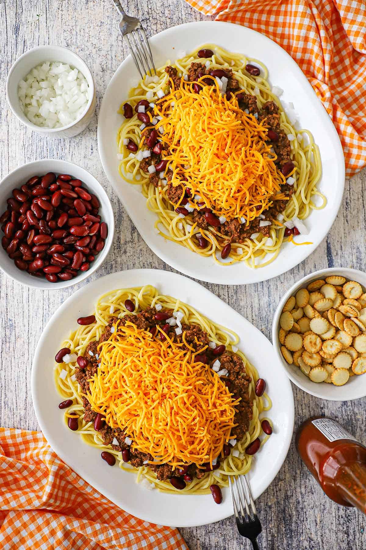 An overhead view of two large oval dinner plates filled with heaping servings of Cincinnati chili served over spaghetti, beans, onions, and topped with shredded cheddar cheese.