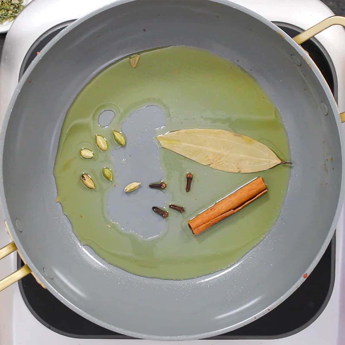 A large non-skillet filled with simmering ghee with a cinnamon stick, an Indian bay leaf, cloves, and green cardamom pods.
