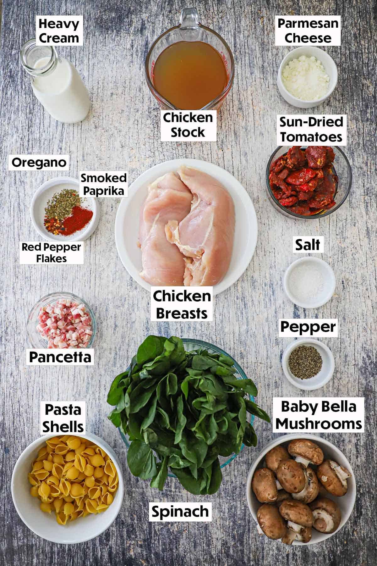 An arrangement of ingredients for One-Pot Marry Me Chicken Pasta on a wooden background including chicken breasts, cream, cheese, sun-dried tomatoes, spinach, pancetta, pasta, mushrooms, and seasonings.