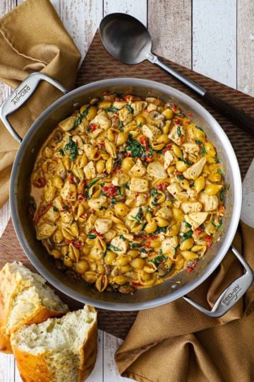 An overhead view of a large filled with One-Pot Marry Me Chicken Pasta including cooked pasta shells, bites of chicken, sun-dried tomatoes, wilted spinach, all in a creamy Tuscan sauce.