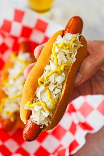 A person holding a Coney Island hot dog in his hand with the frankfurter nestled into the bun and topped with sauerkraut, chopped onions, and a zig zag of deli-style mustard.
