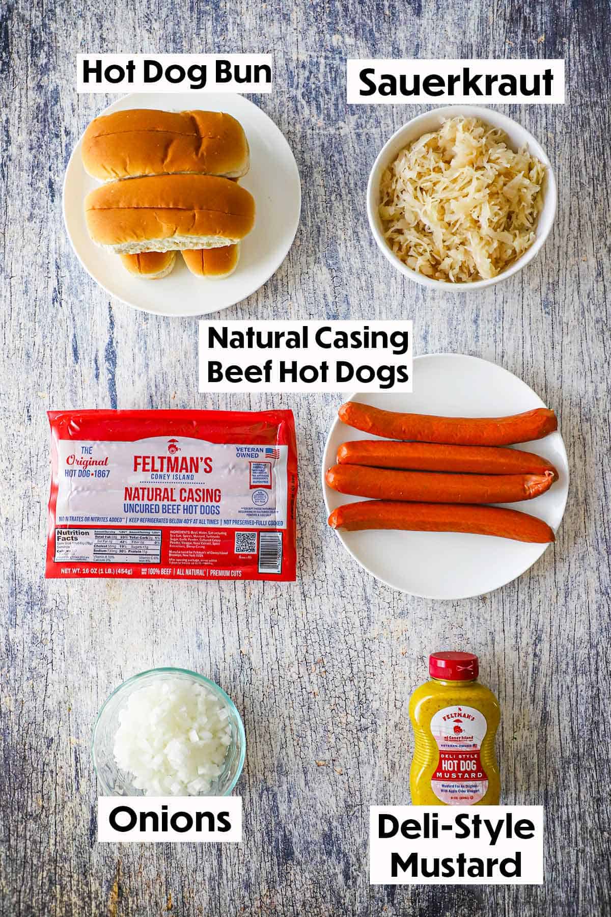 An arrangement of ingredients for Coney Island hot dogs on a wooden grey background including a package of Feltman's Natural Casing hot dogs, a white plate of hot dogs, buns, sauerkraut, chopped onions, and a bottle of deli-style mustard.