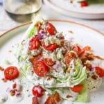 A steakhouse wedge salad with homemade blue cheese on a white dinner plate with a stemless glass of white wine in the background.