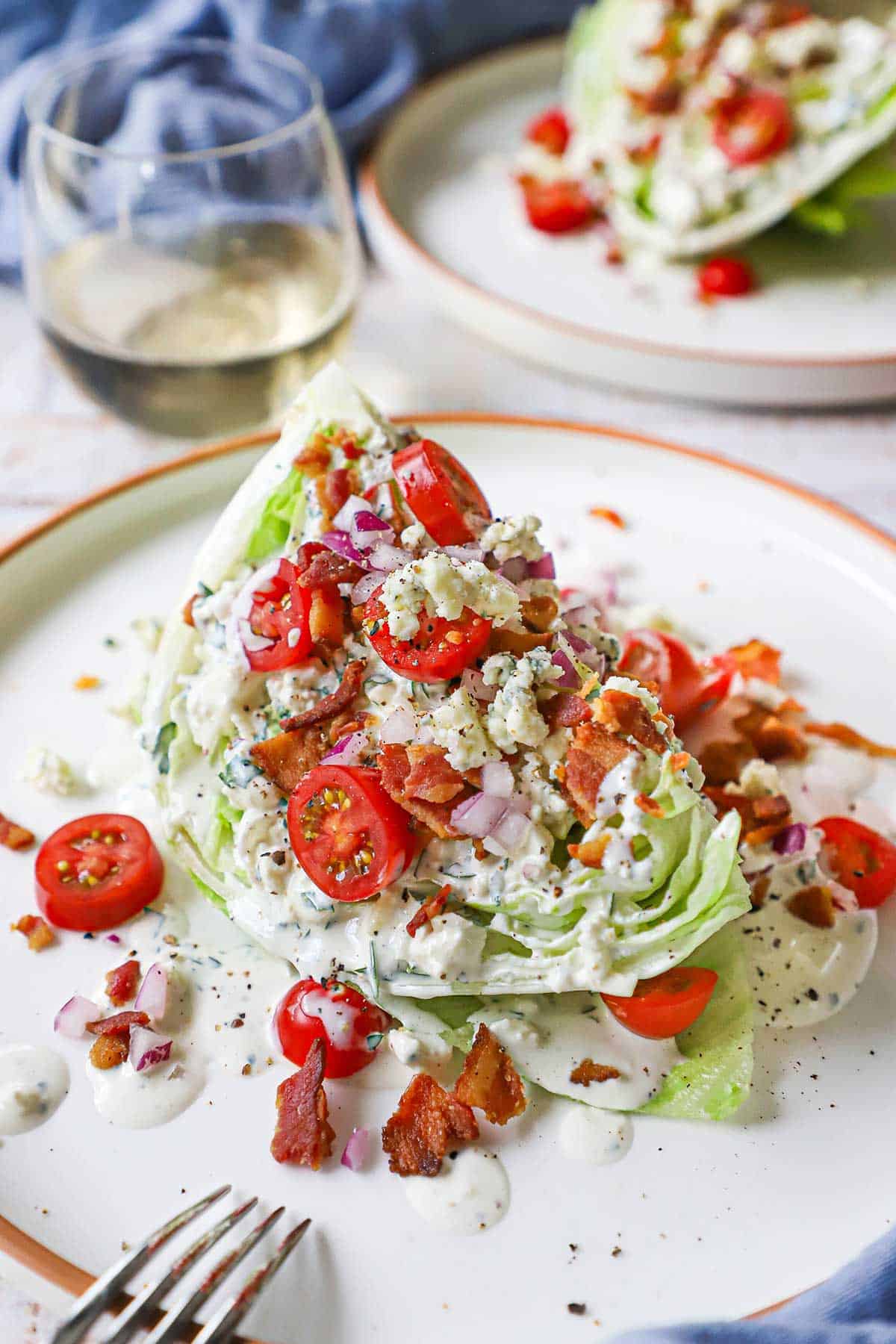 A steakhouse wedge salad with homemade blue cheese on a white dinner plate with a stemless glass of white wine in the background.
