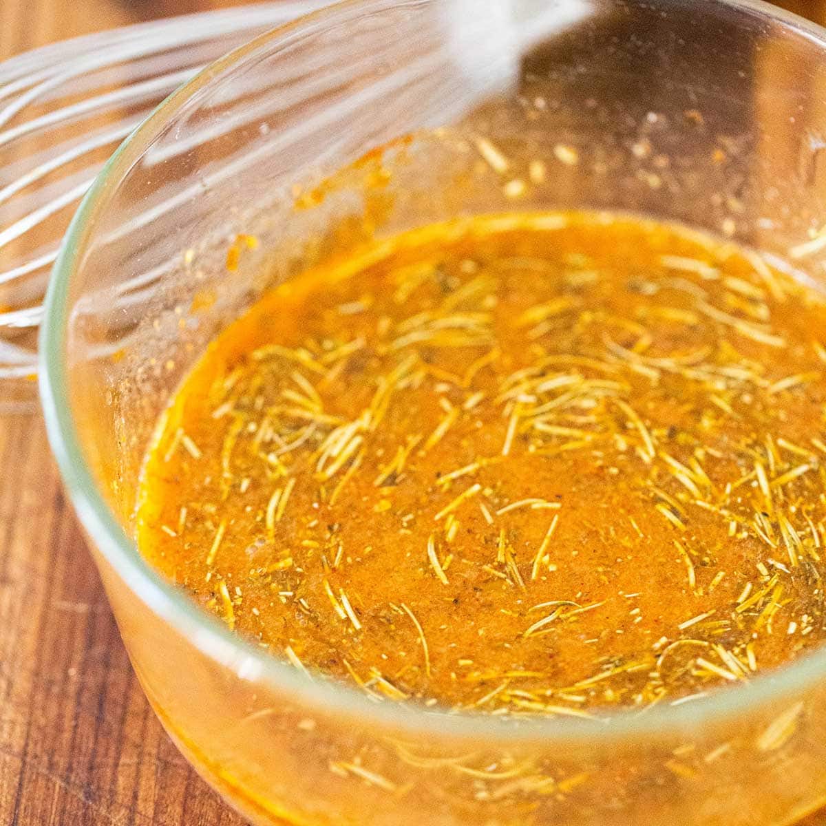 A close-up view of a butter, citrus, and herb marinade in a small glass bowl with a whisk sitting next to it.