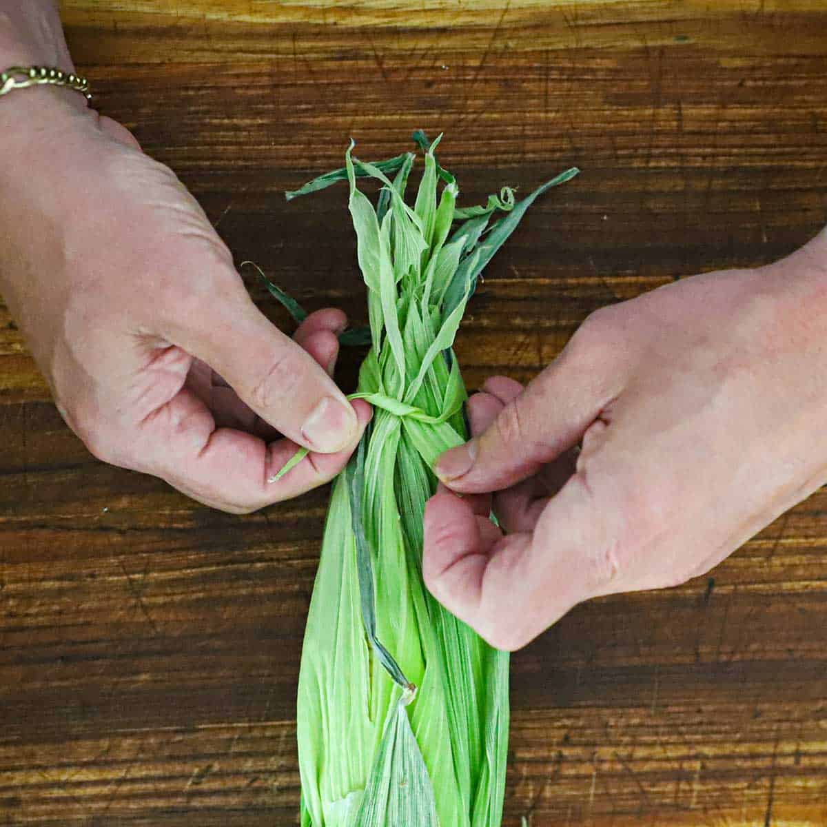 A person using a thin strip of a corn husk to tie together the entire husk at the end of an ear of corn on a wooden cutting board.