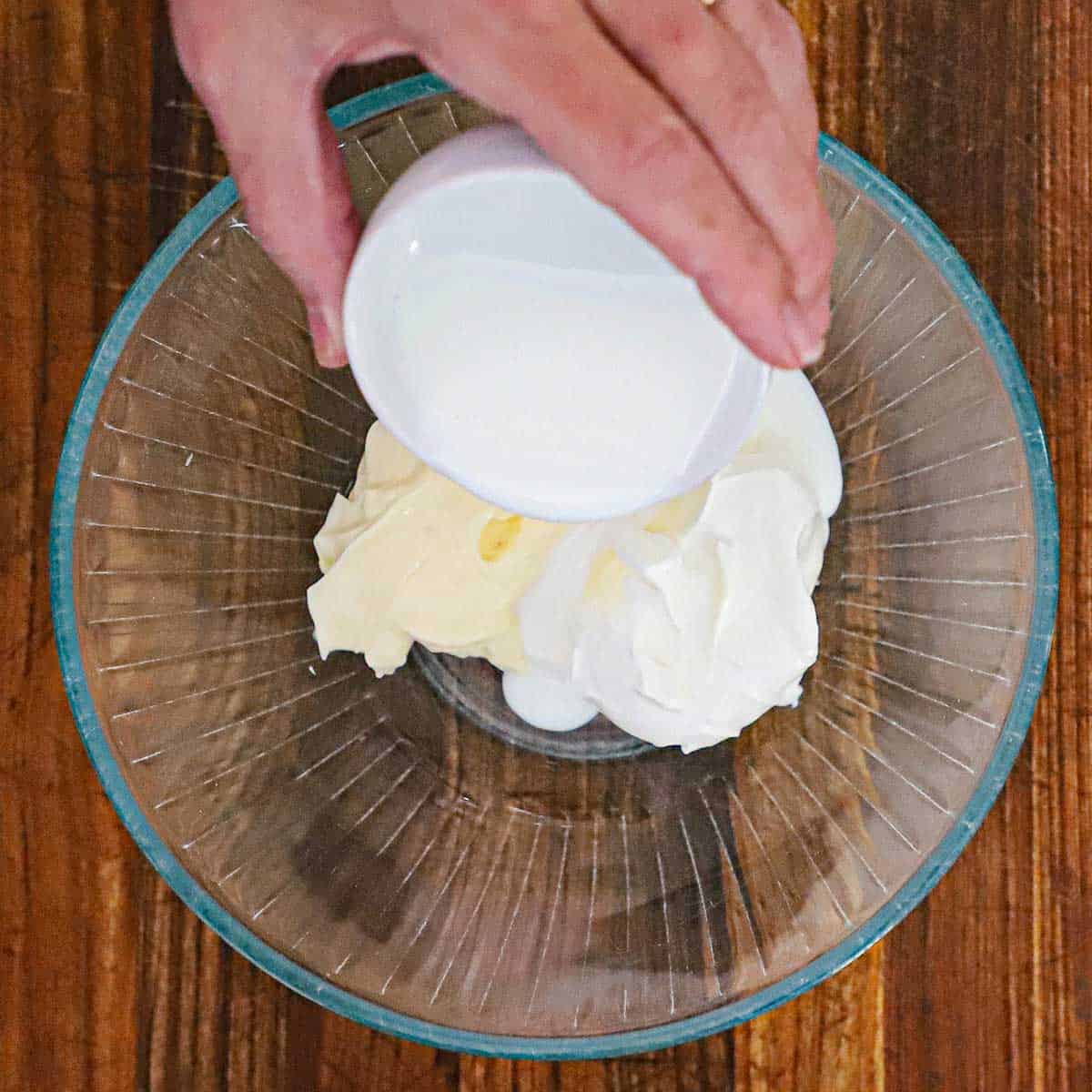 A person pouring buttermilk from a small white bowl into a glass bowl that contains small mounds of mayonnaise and sour cream.