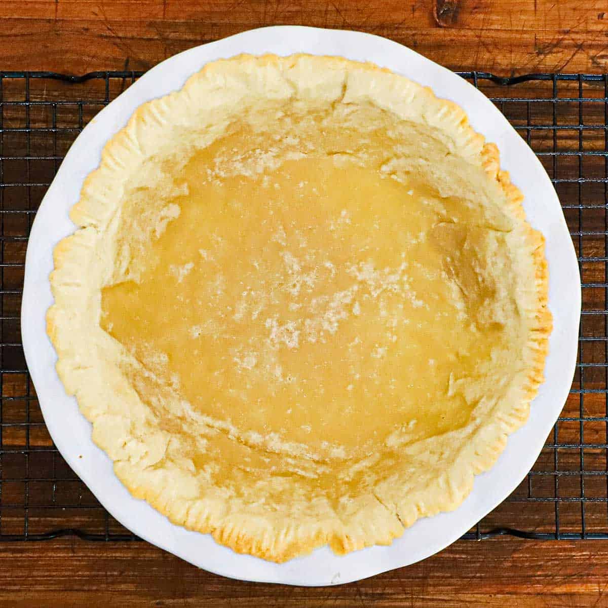 A partially baked pie crust in a white ceramic pie dish resting on a baking rack on a wooden cutting board.