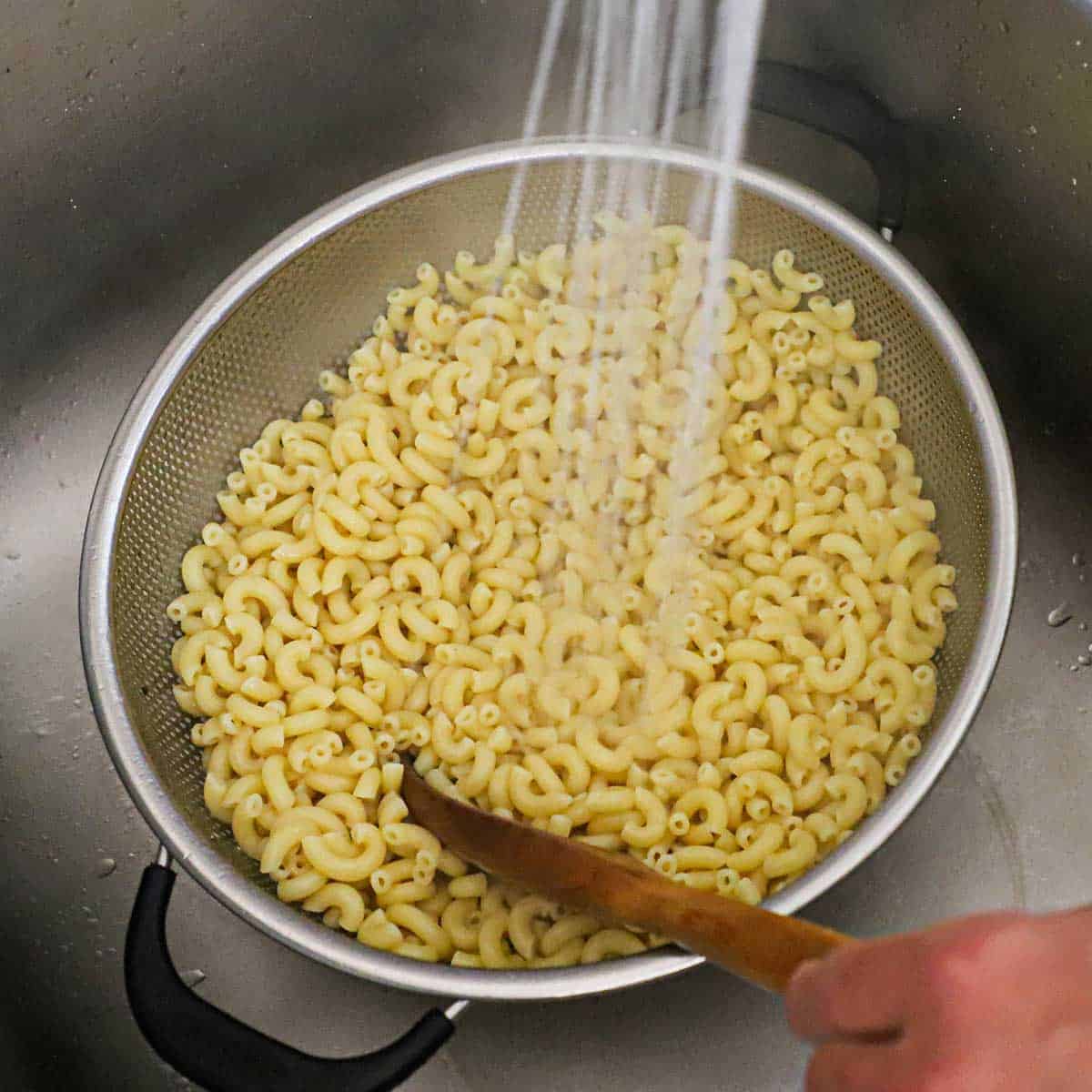 A person using a sink faucet to spray tap water over cooked elbow macaroni that is in a colander in a kitchen sink.