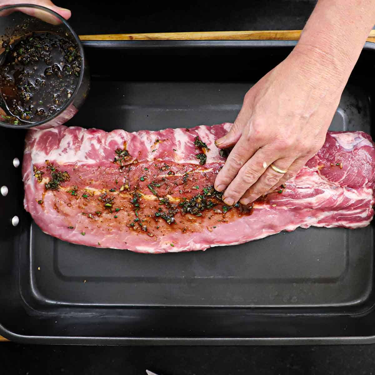 A person using his fingers to press a balsamic and herbs marinade into an uncooked rack of baby back ribs that is resting in a large black roasting pan.