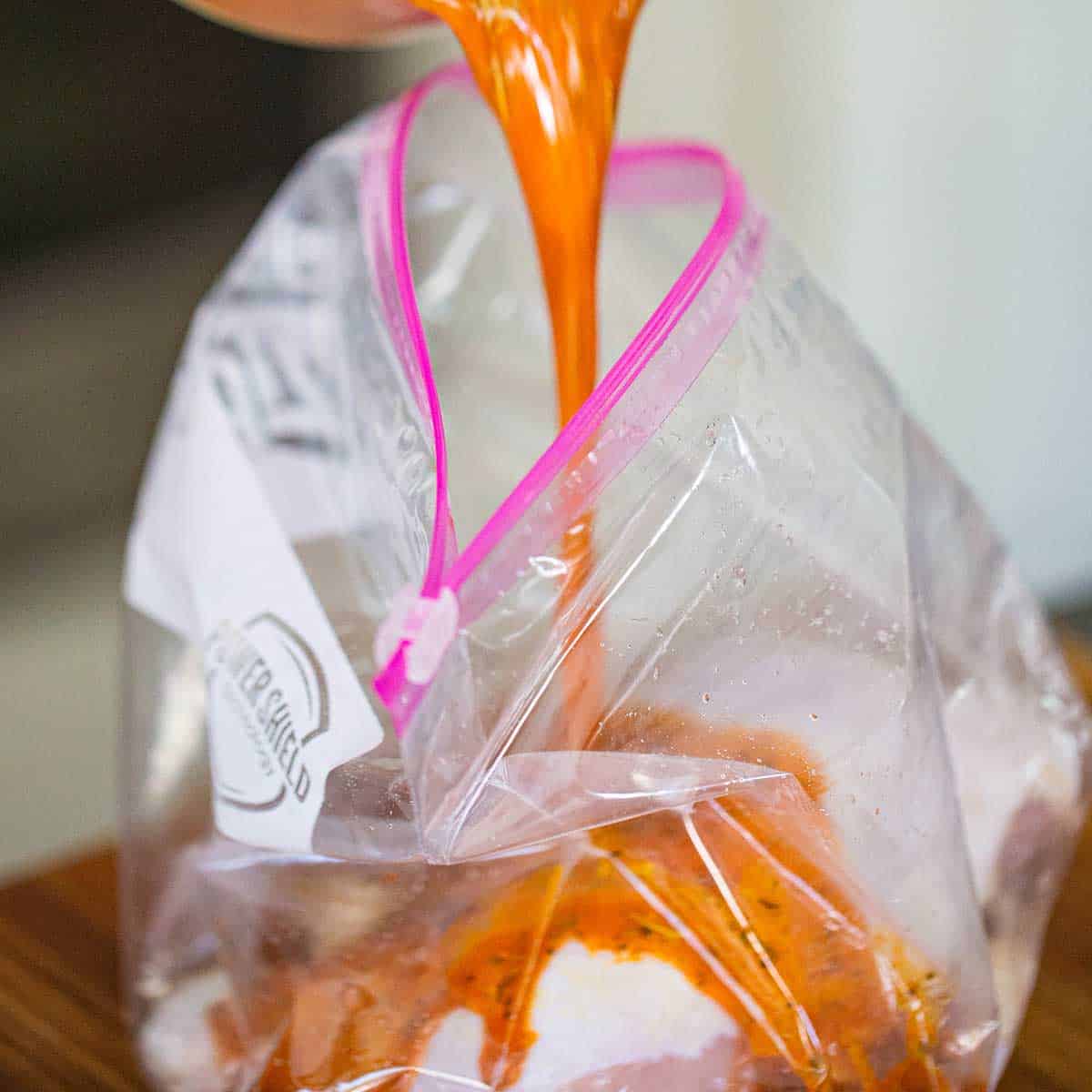 A person pouring a marinade into a large plastic baggie filled with raw chicken leg quarters.