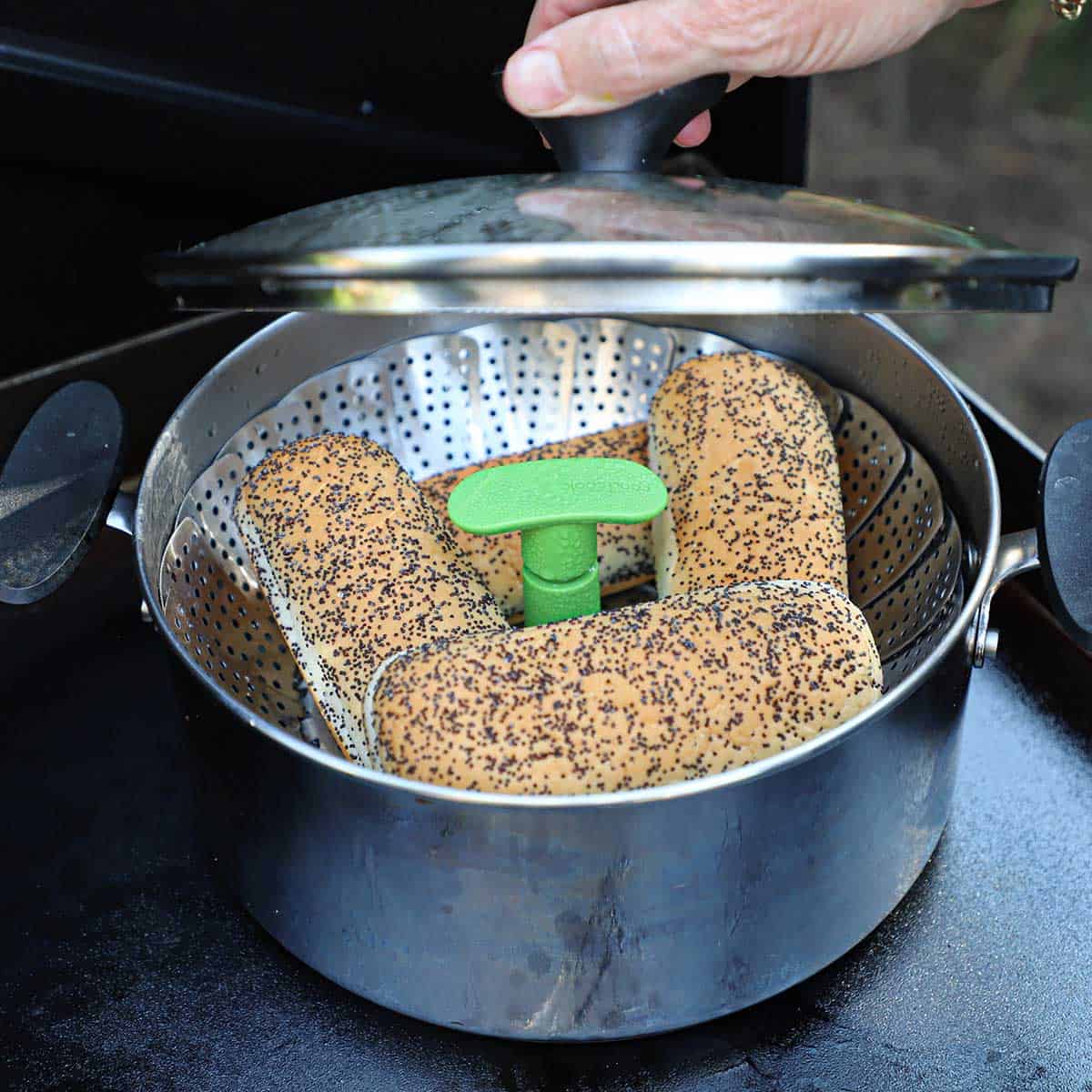 A person lifting a lid from a pot that is lined with a steam basket and is holding four poppyseed hamburger buns.