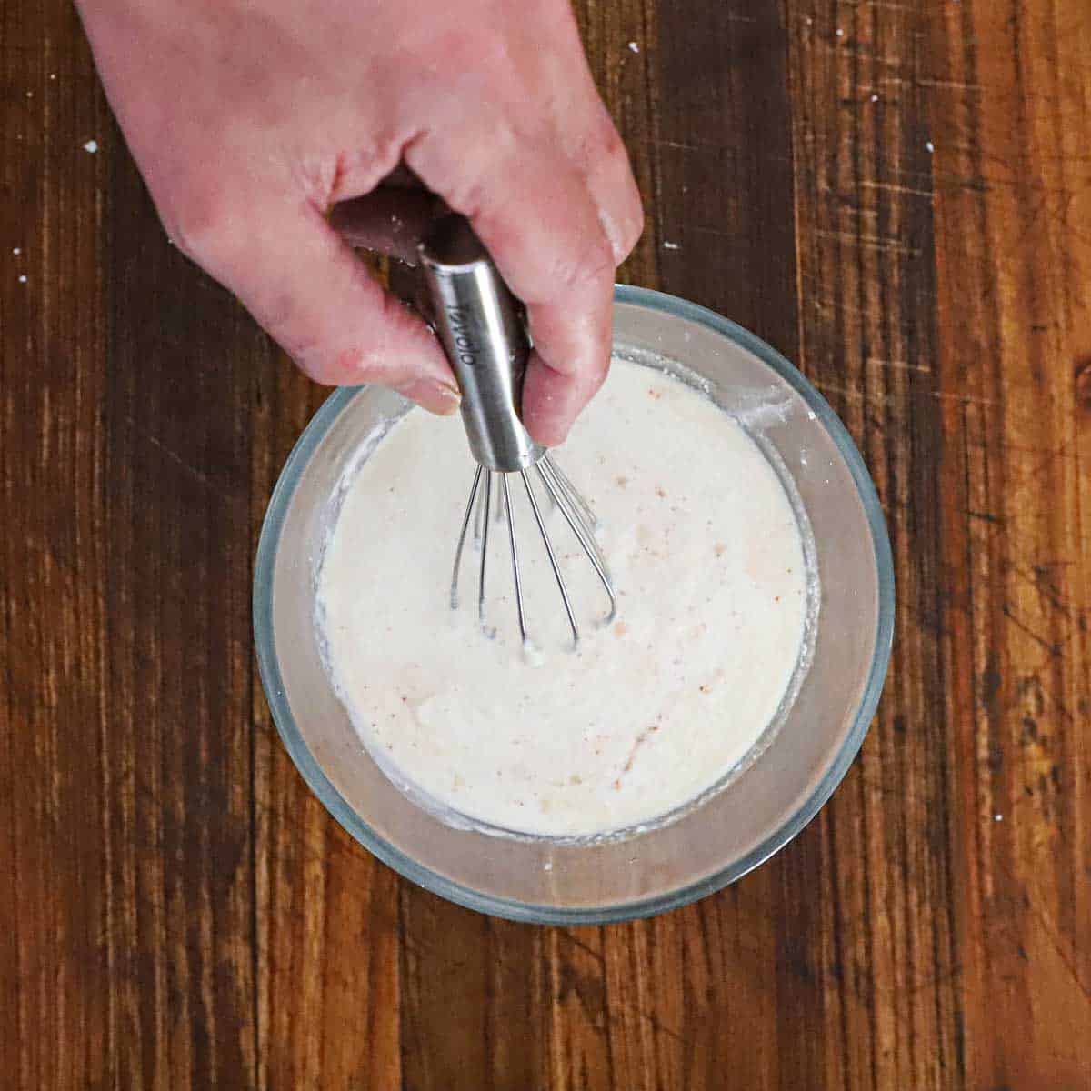 A person whisking a cream and Cotija cheese mixture in a small glass bowl on a wooden cutting board.