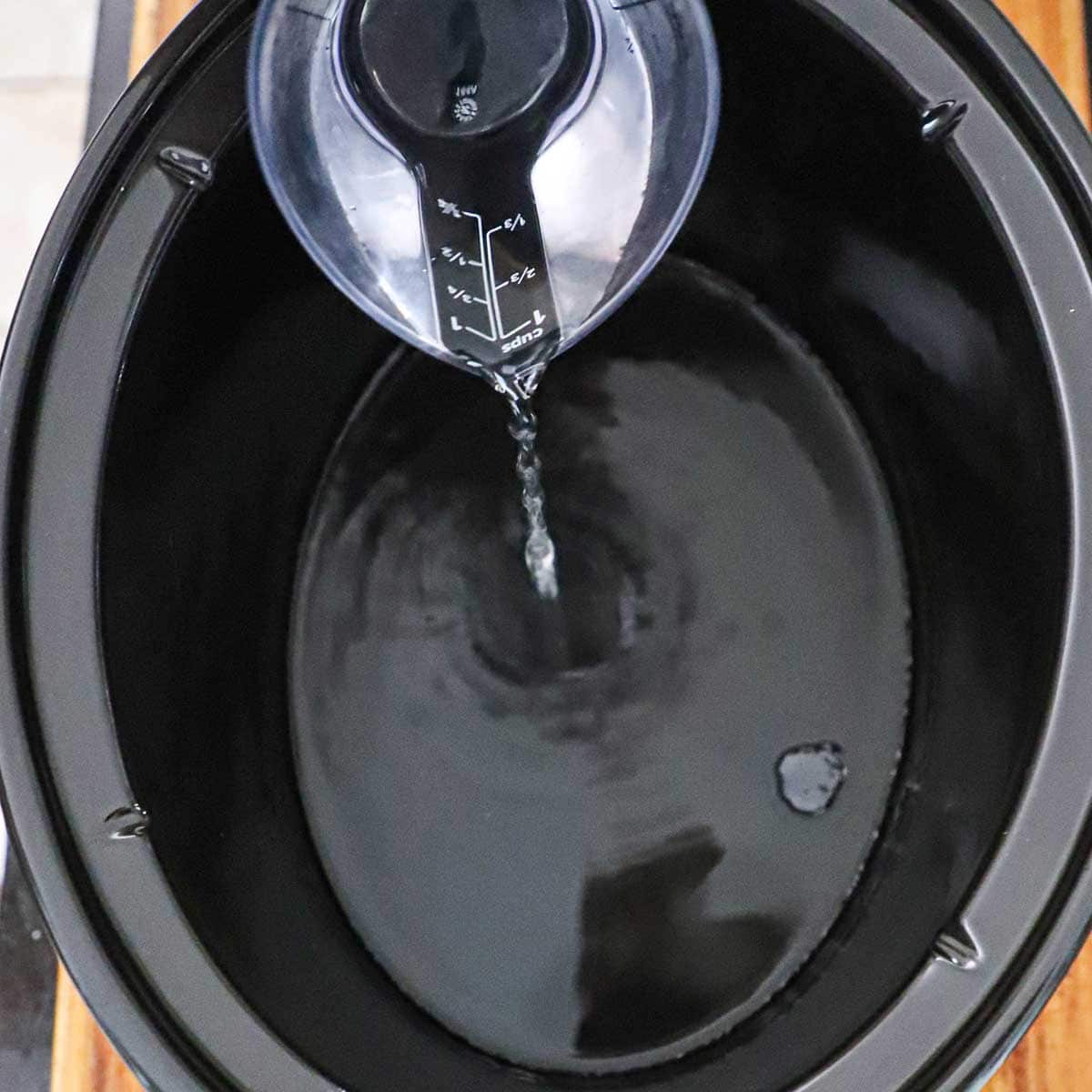 A person pouring water from a 1-cup plastic measuring cup into the base of a black slow cooker.