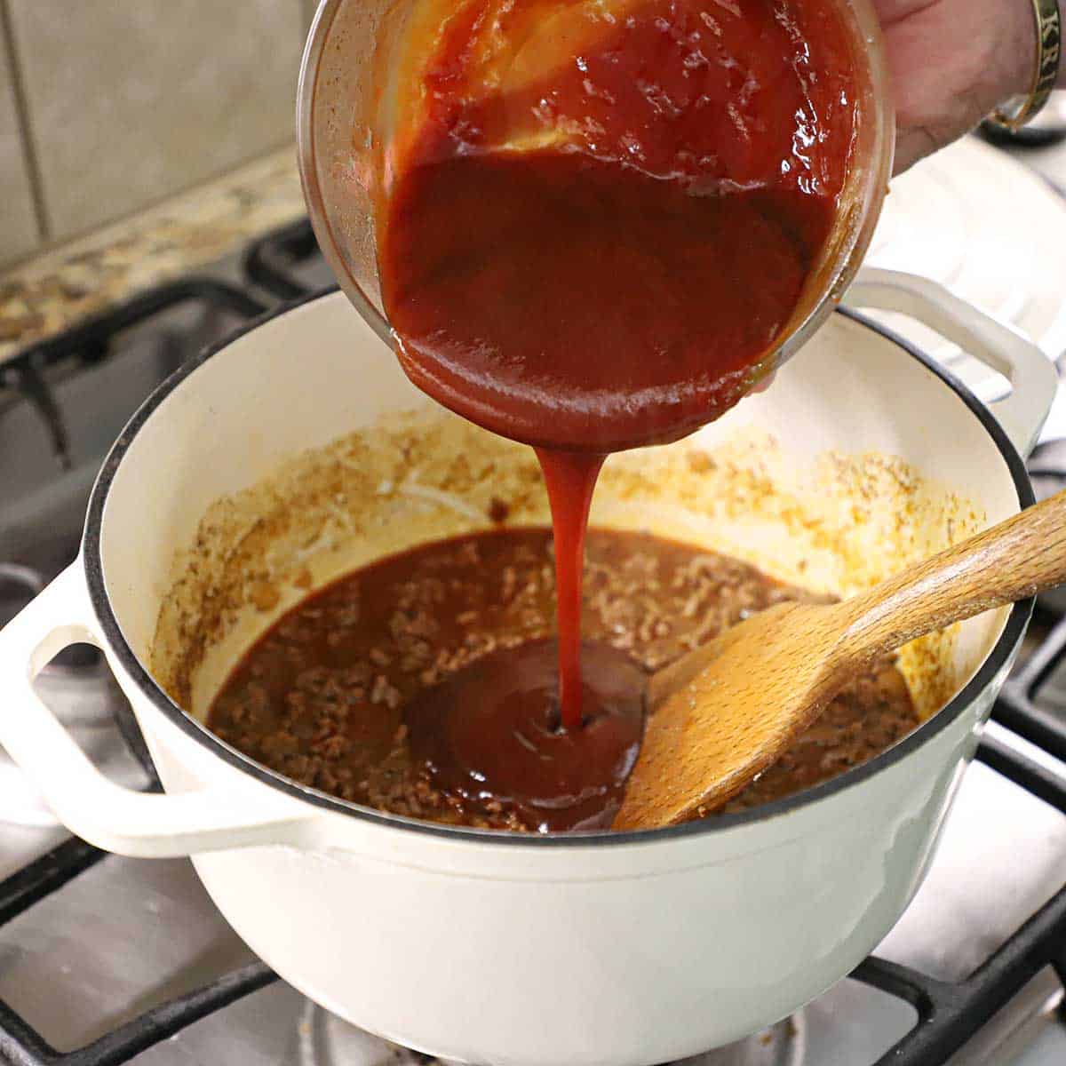 A person pouring a tomato and brown sugar mixture into a pot of simmering meat sauce in a white pot on a gas stove.