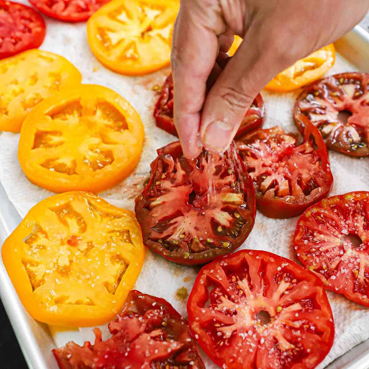 A person sprinkling Kosher salt over slices of red, orange, and yellow heirloom tomatoes that are resting on paper towels in a baking pan.