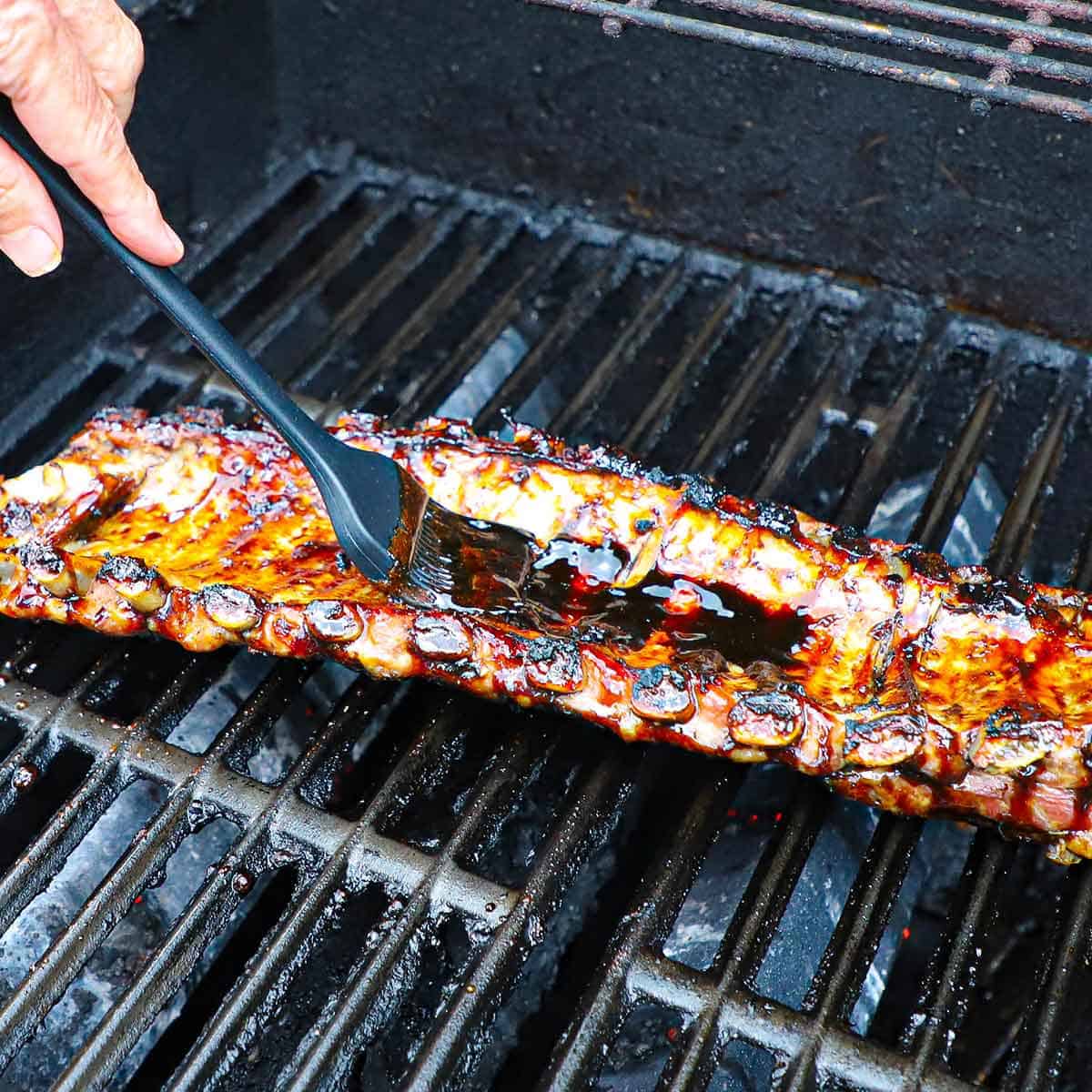 A person using a grill brush to apply a balsamic glaze onto the underside of a rack of baby back ribs being cooked on the grate of a gas grill.