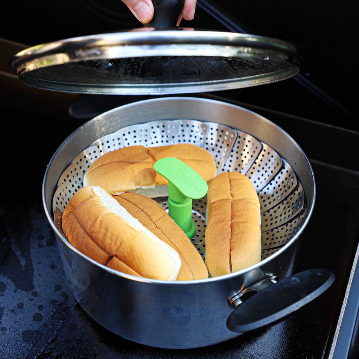Four top-sliced hot dog buns resting on a steamer basket that is in a pot of simmering water on a large black outdoor griddle.