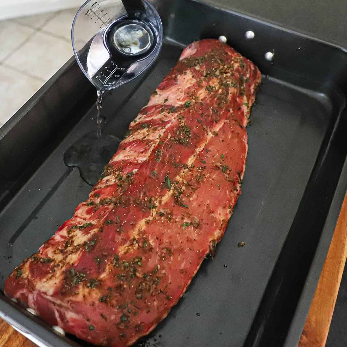 A person pouring water from a measuring cup into a large black roasting pan that is holding a seasoned rack of baby back ribs.