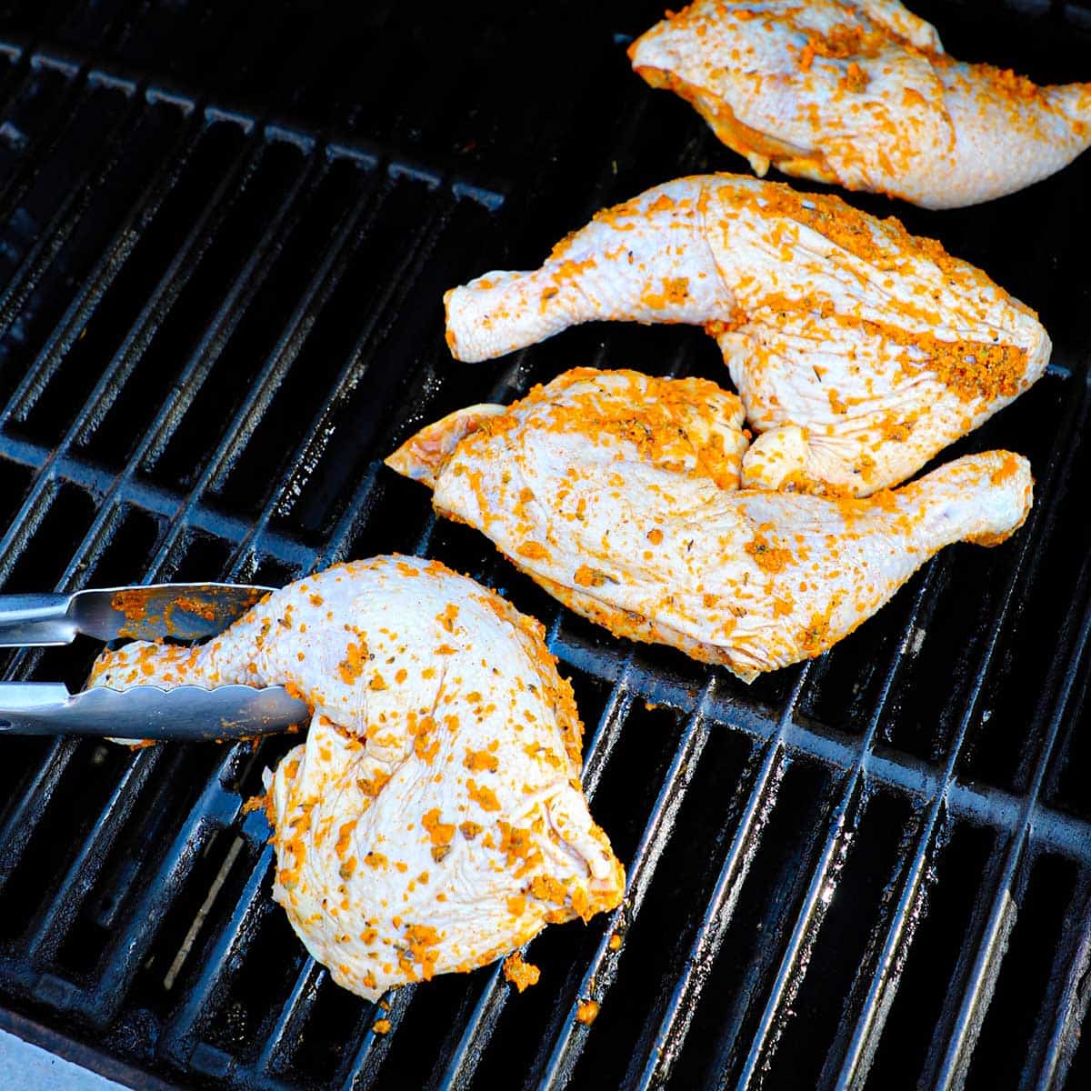 A person using metal tongs to place uncooked chicken leg quarters on the grate of a gas grill.