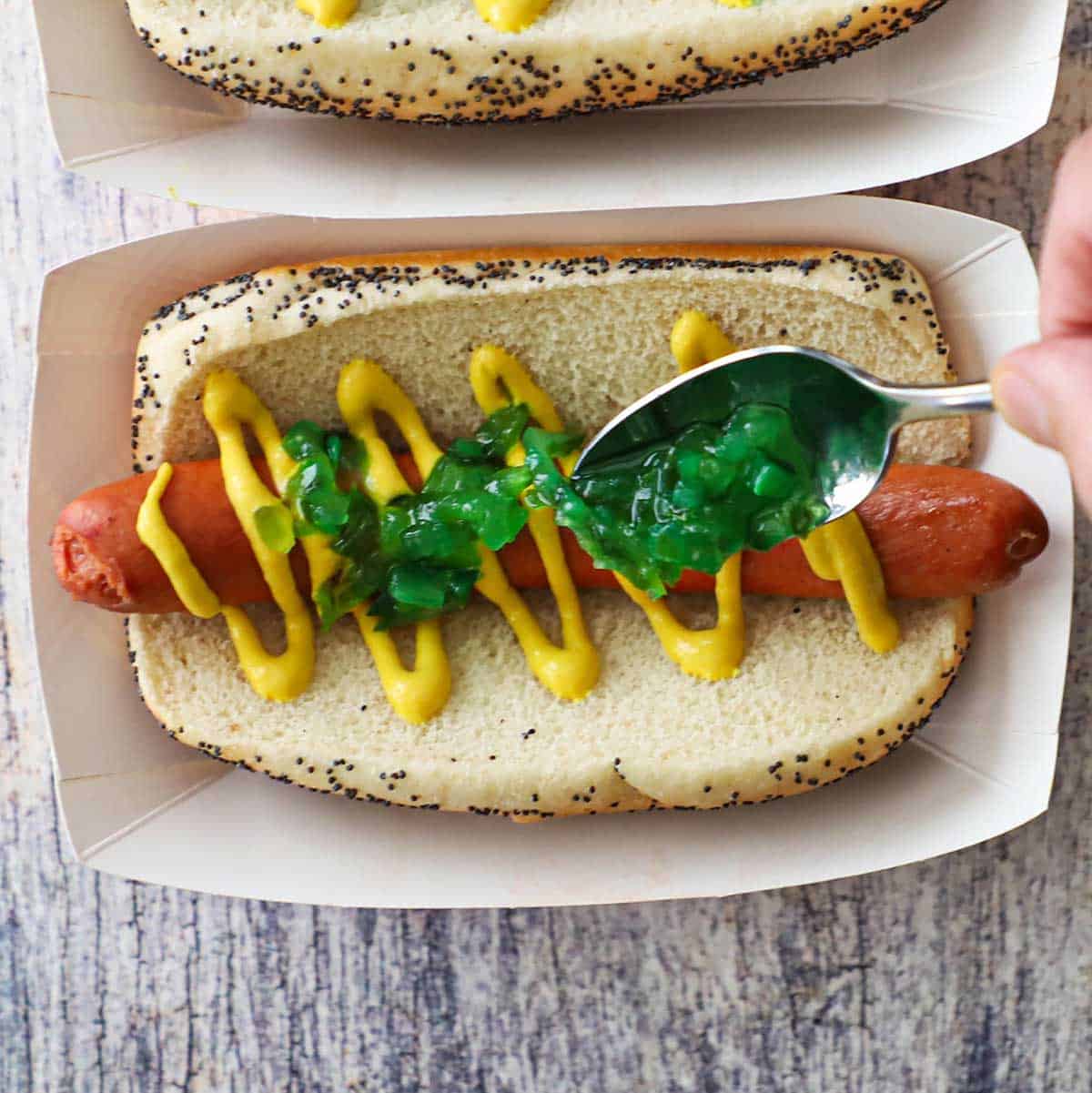 A person using a spoon to add a layer of bright green relish over a hot dog in a poppyseed bun topped with a zig-zag of yellow mustard.