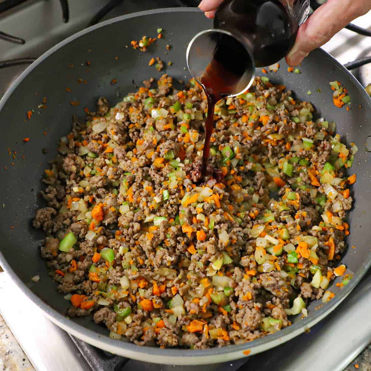 A person pouring red wine from a small carafe into a large skillet filled with cooked beef and Italian sausage with chopped onion, celery, and carrots.