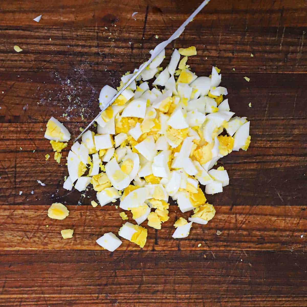 A person using a large knife to chop hard boiled eggs on a large wooden cutting board.