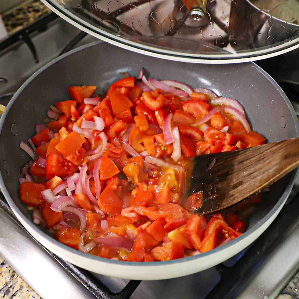 A large non-stick skillet that is filled with simmering chopped tomatoes with softened strips of red onion with a wooden spatula inserted into the side.