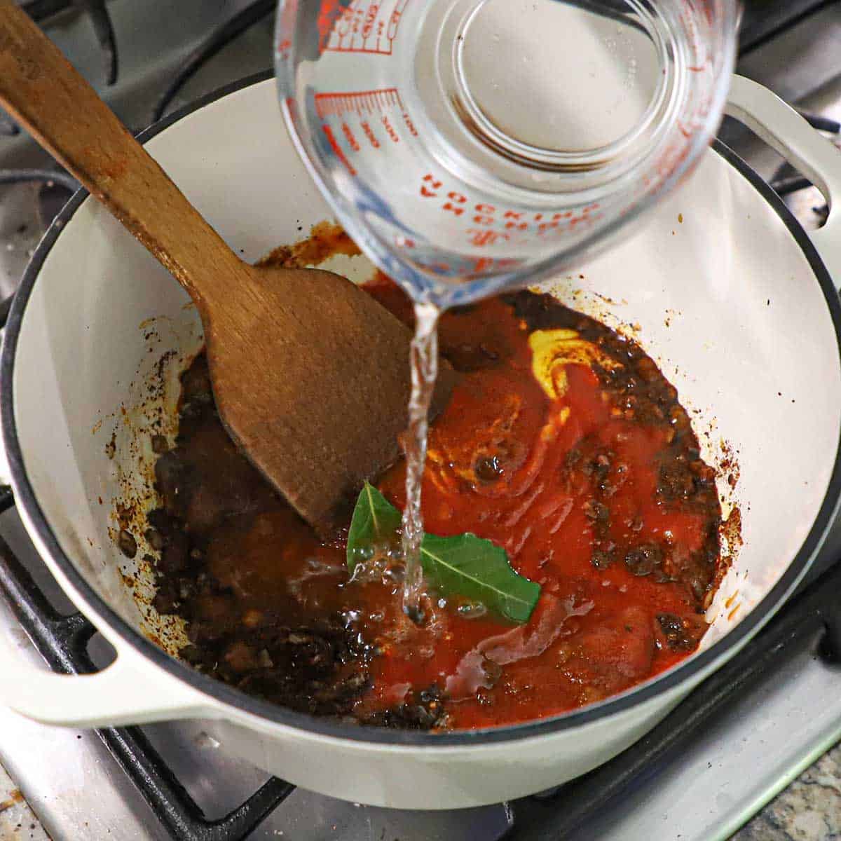 A person pouring water from a glass measuring cup into a pot filled with seasoned sautéd onions, tomato sauce, yellow mustard, and two bay leaves.