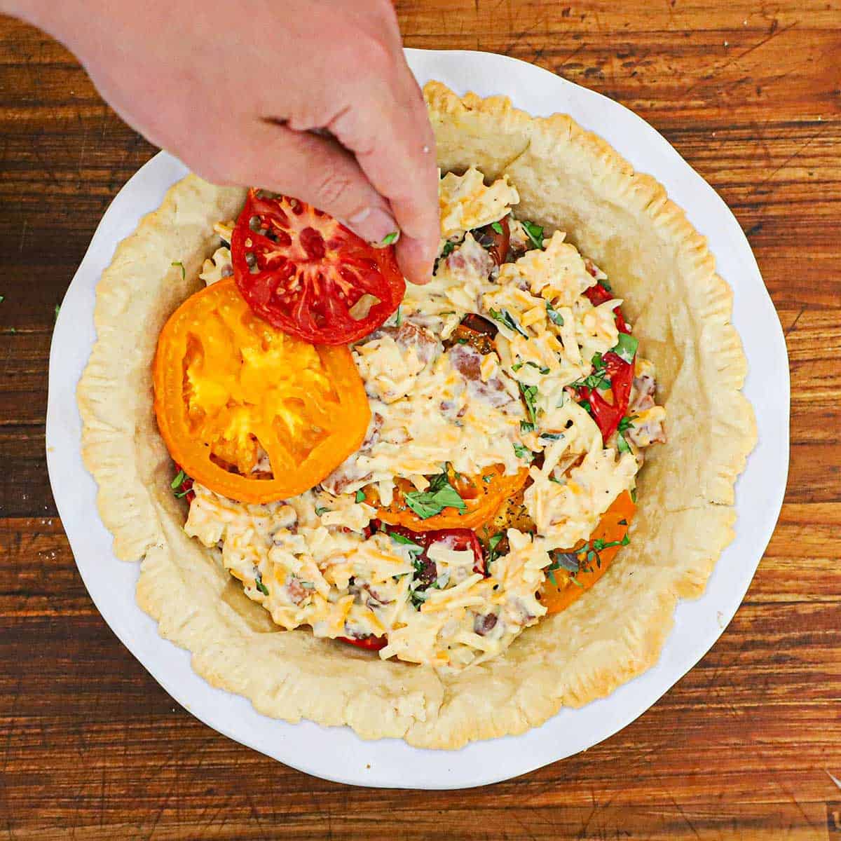 A person adding sliced heirloom tomatoes onto layers of a mayonnaise and cheese mixture, herbs, and more heirloom tomatoes in a pie dish lined with a partially baked pie crust.