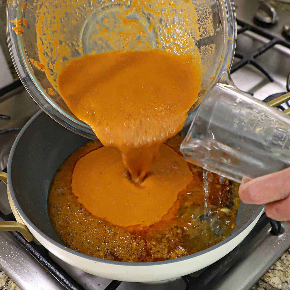 A person pouring butter masala gravy from a glass bowl along with water from a measuring cup into a large skillet filled with simmering masala sauce.