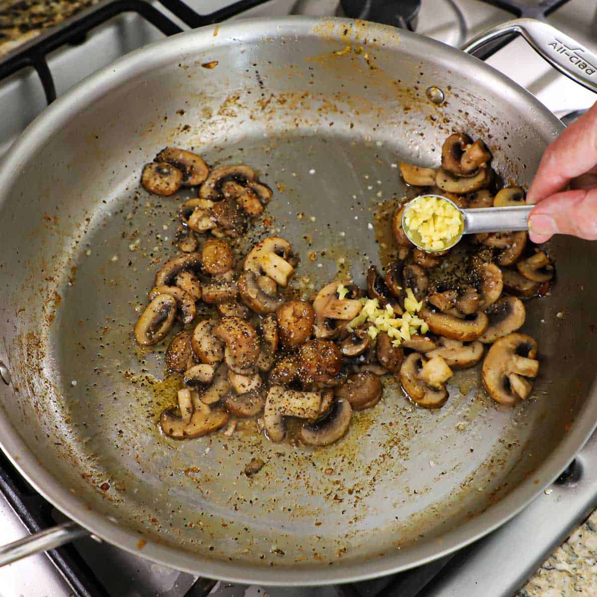 A person transferring minced garlic from a teaspoon into a skillet that is filled with sautéed mushrooms that have been seasoned with salt, pepper, and dried oregano.
