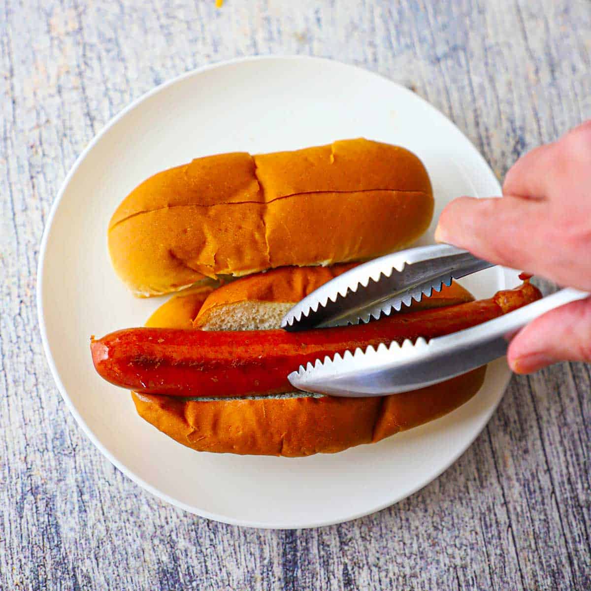 A person using a pair of tongs to place a natural casing beef frankfurter into a top-sliced bun on a white plate.