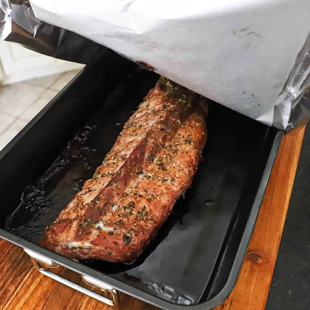A person pulling back a large sheet of aluminum foil from large black roasting pan that has a roasted rack of baby back ribs in it.