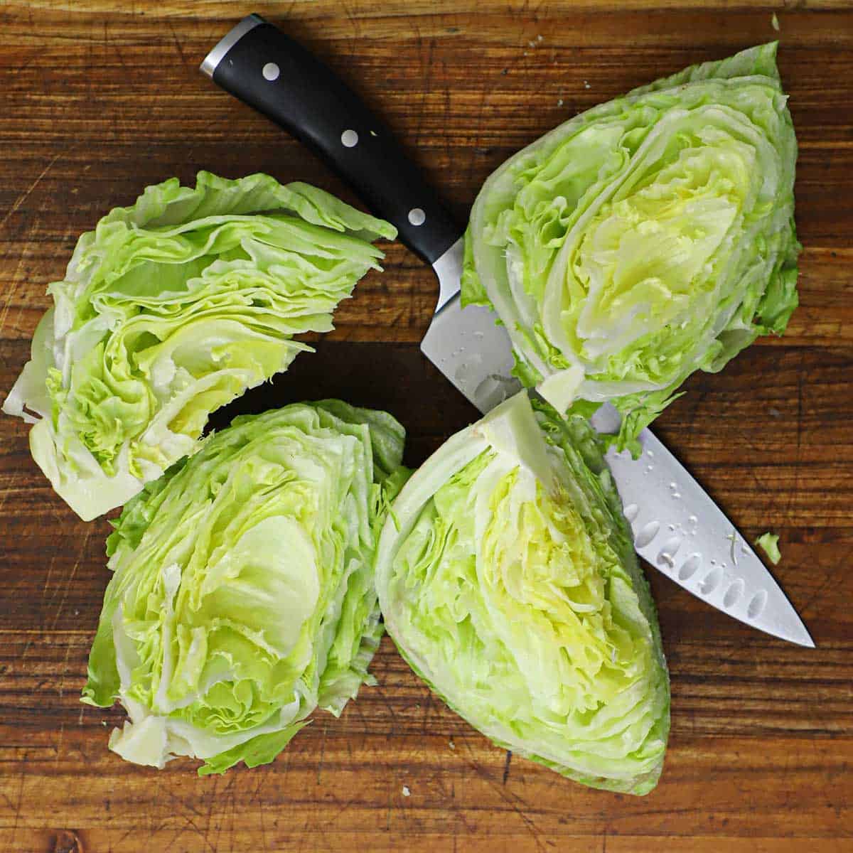 A head of iceberg lettuce that has been cut into four wedges and are resting on a large chef's knife on a wooden cutting board.