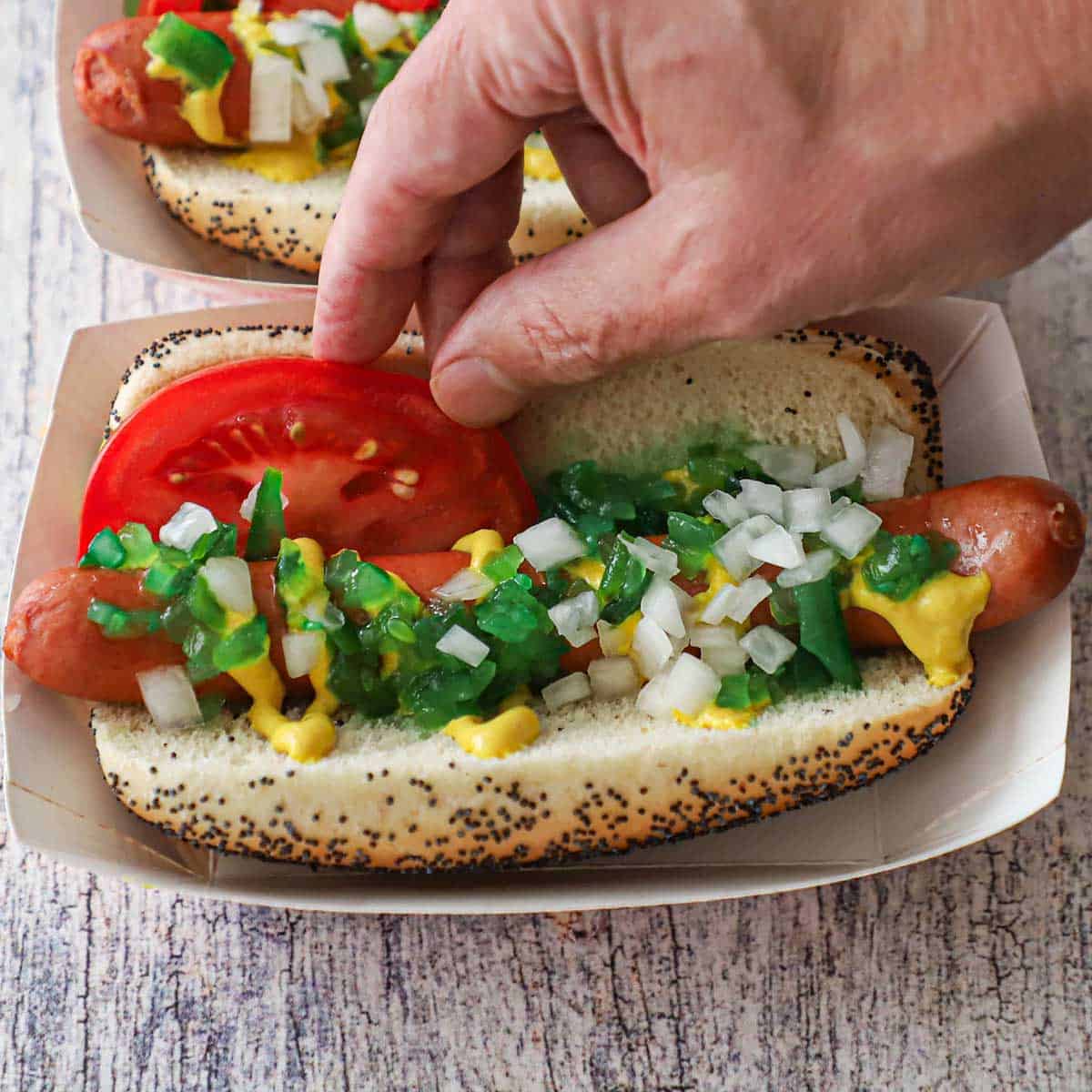 A person nestling a tomato wedge beside a hot dog that is in a poppyseed bun and is topped with yellow mustard, green relish, and chopped onions.