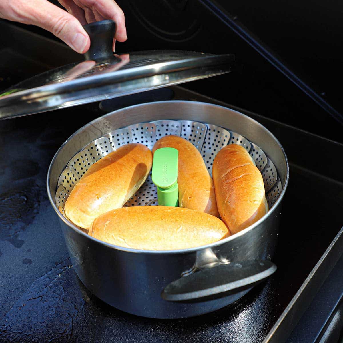 A person removing a lid from a pot that has been fitted with a steam basket with four hot dog bun resting in the basket.