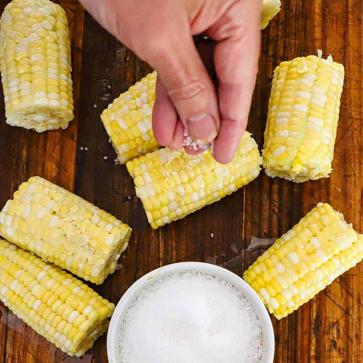 A person using his fingers to sprinkle Kosher salt over uncooked ears of corn the have been cut in half and are resting on a wooden cutting board.