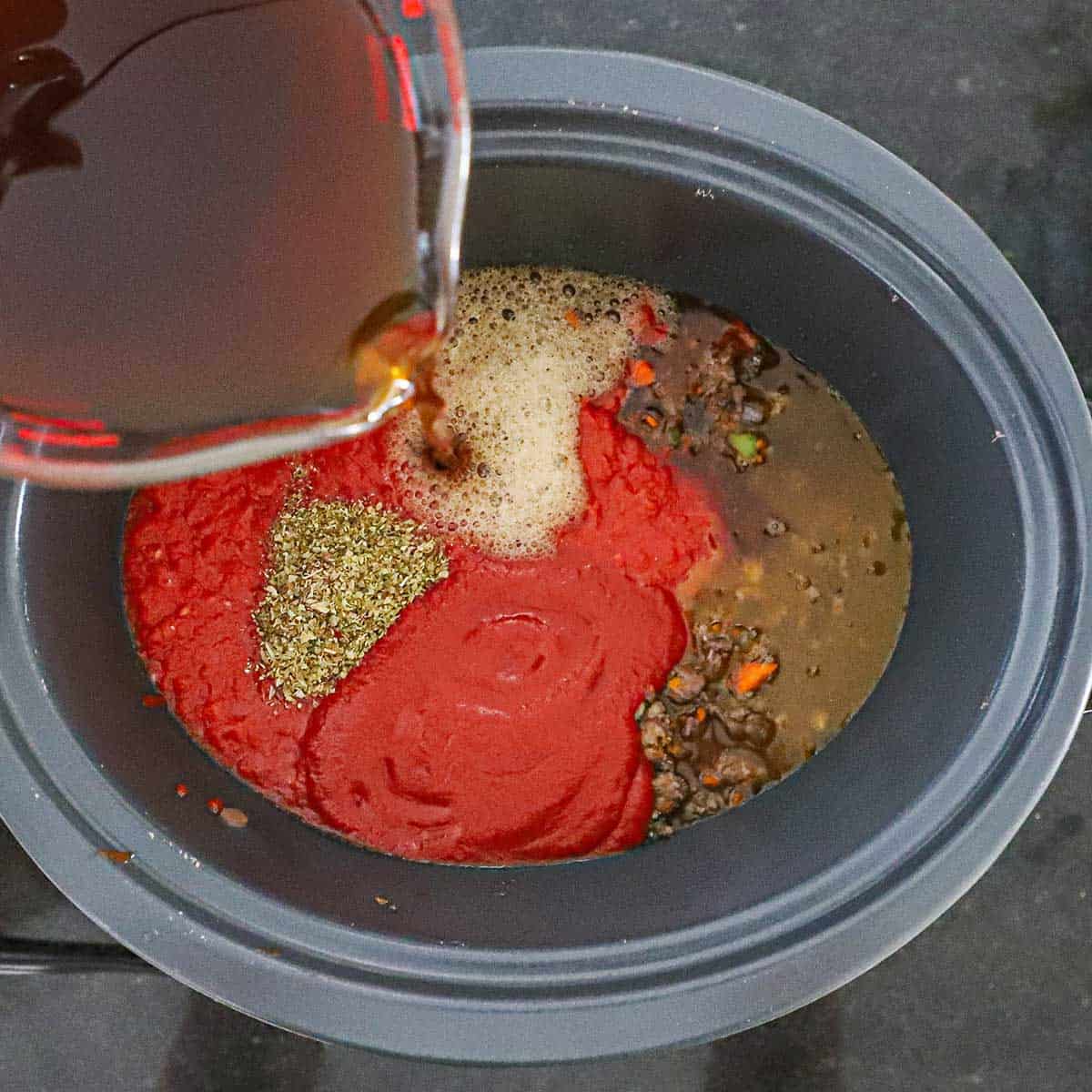 A person pouring beef broth from a large glass measuring cup into a slow cooker filled with crushed tomatoes, dried seasonings, and ground beef with sautéed vegetables.