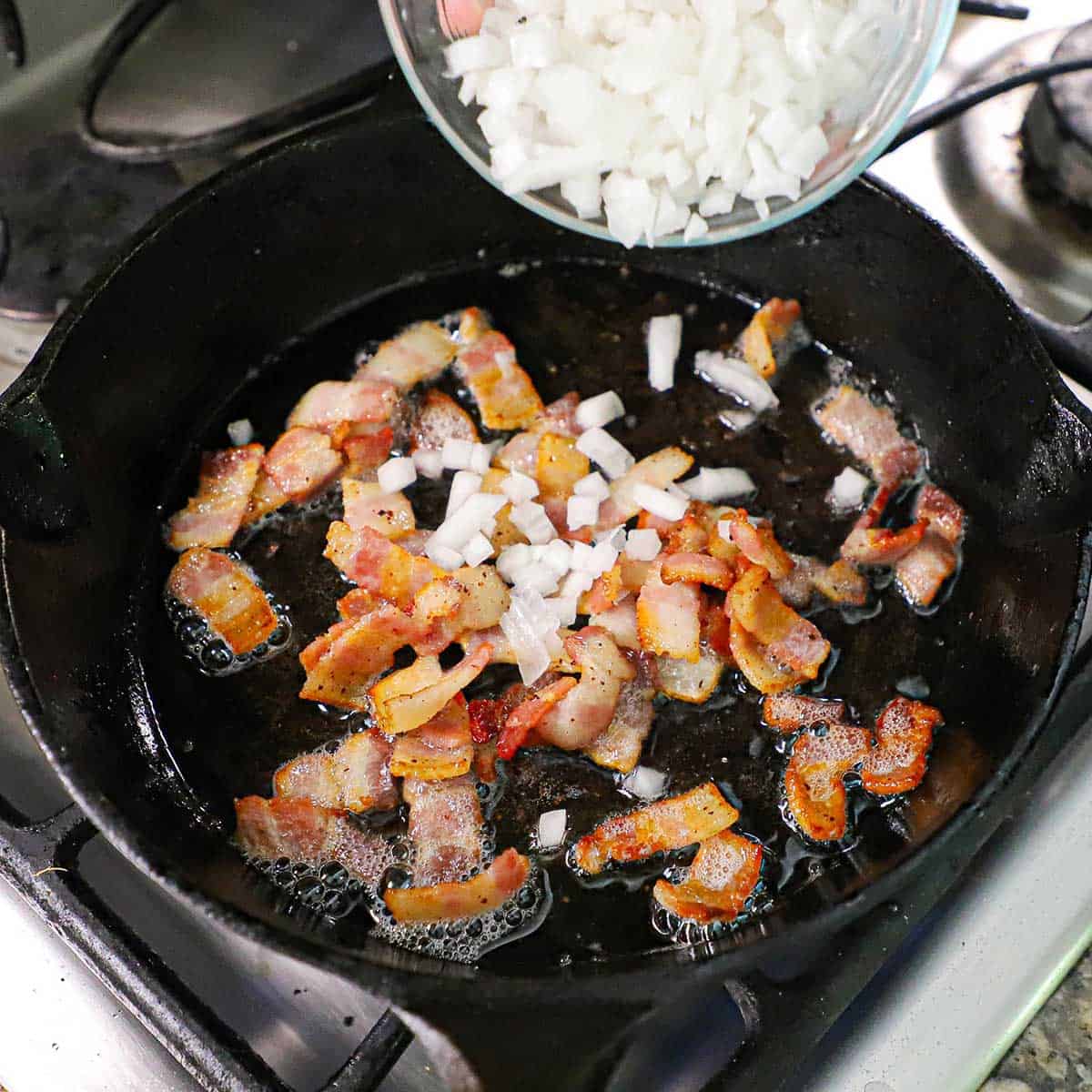 A person dumping chopped onions from a small glass bowl into a cast-iron skillet that is filled with crispy chopped bacon that is simmering in bacon grease.