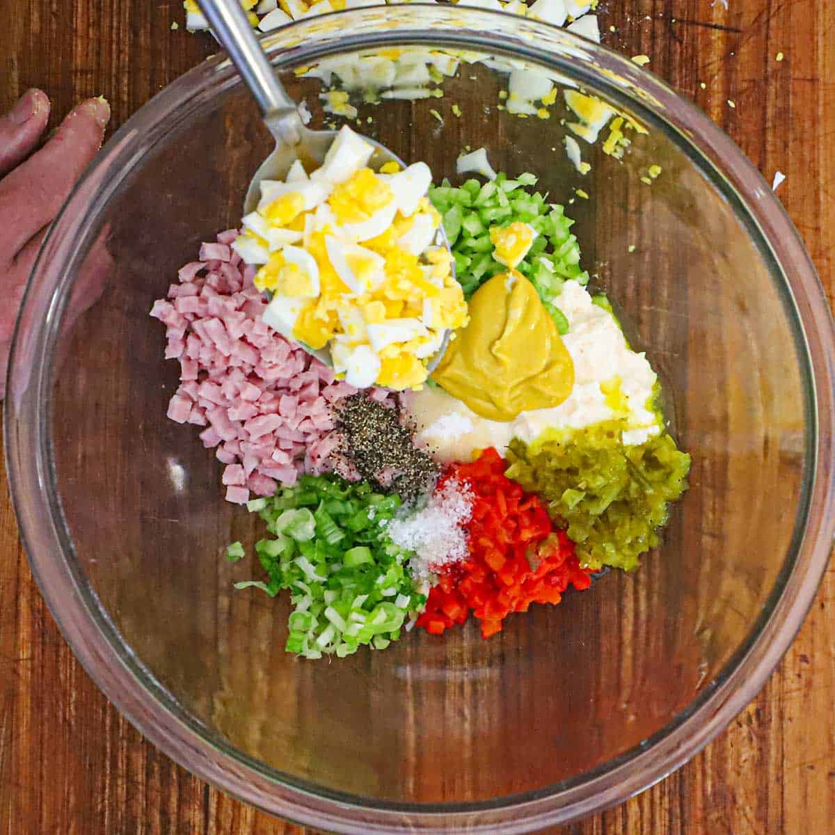 A large glass bowl filled with hopped ham, chopped scallion, pimentos, relish, mustard, mayonnaise, celery, and spoonful of chopped hard boiled eggs.
