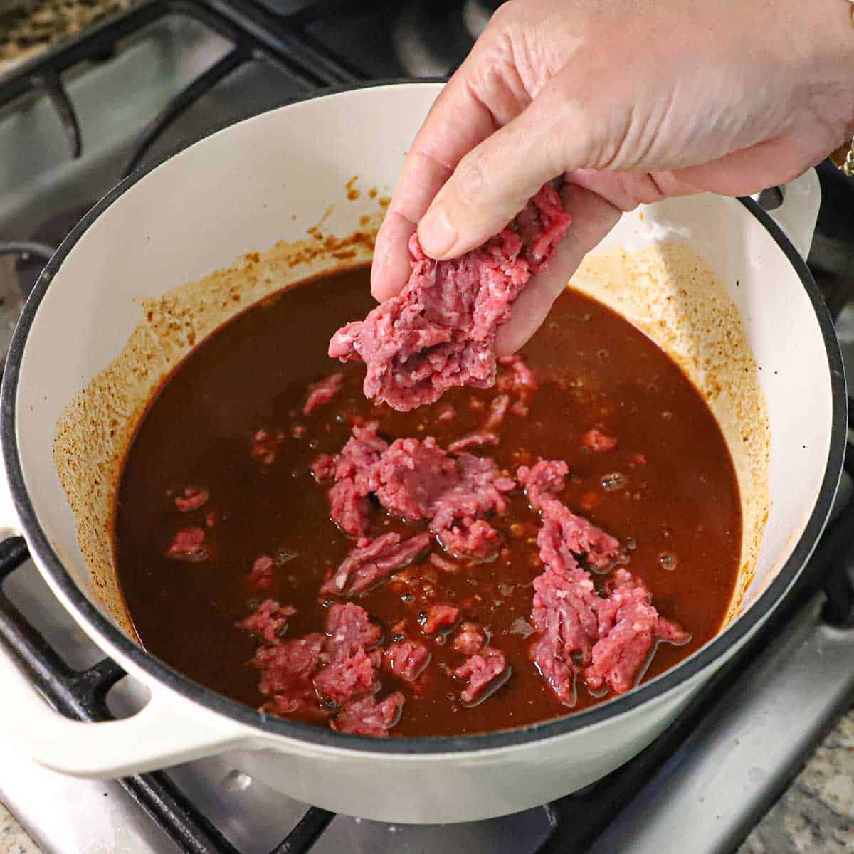 A person using his hand to crumble uncooked ground beef into a pot of a simmering tomato-based sauce on a gas stove.