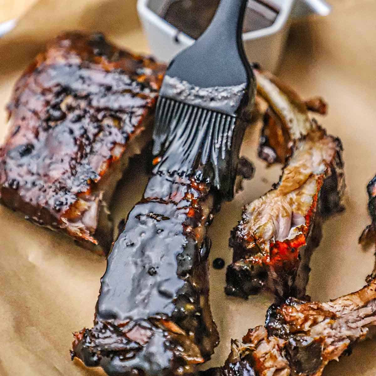 A person using a grill brush to apply more balsamic glaze to an individual baby back rib that sitting on brown paper next to other balsamic glazed ribs.