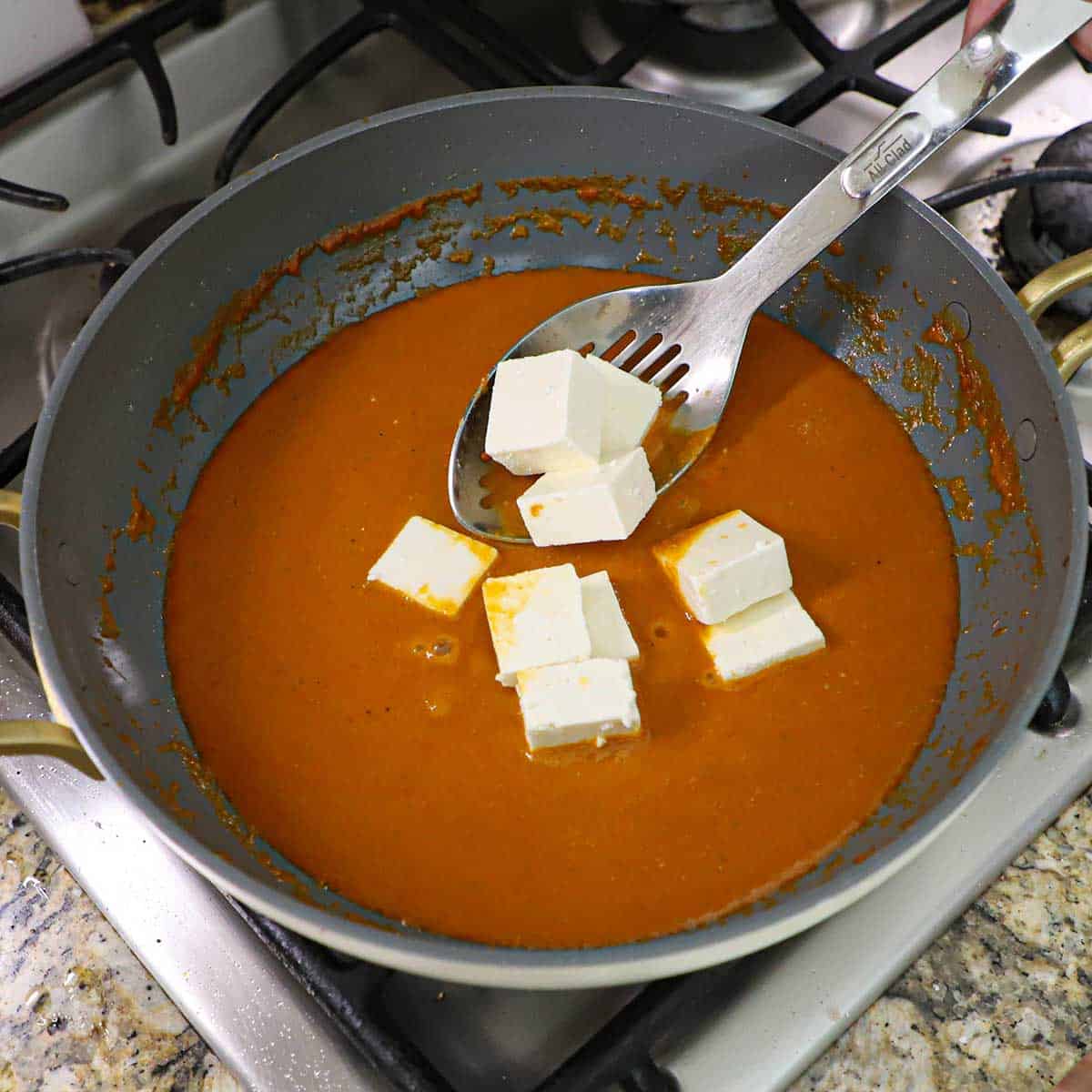 A person using a slotted spoon to lower bite-sized pieces of paneer into a skillet filled with a simmering butter masala sauce.