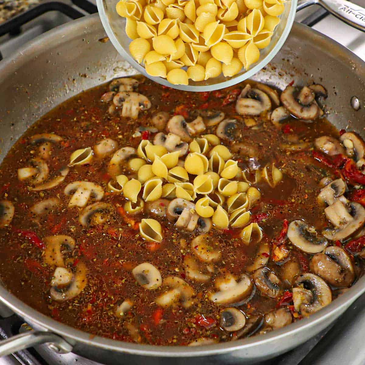 A person transferring dried pasta shells into a large skillet that is filled with a simmering broth that include sautéed slices of mushroom and seasonings.