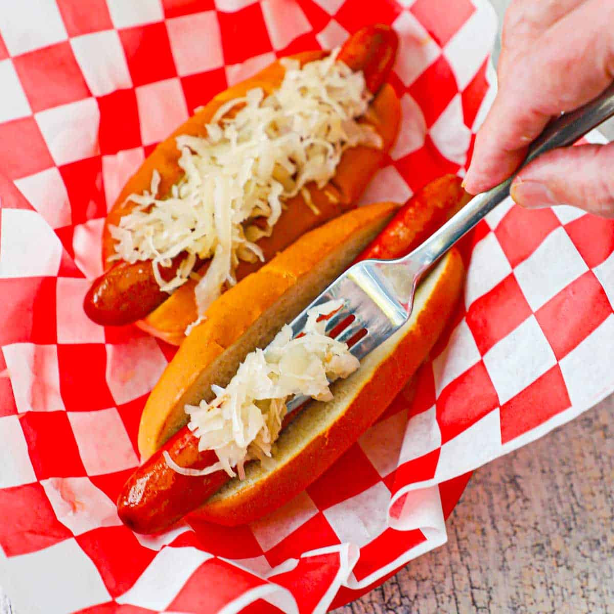 A person using a fork to add a mound of sauerkraut over a beef frankfurter that is resting in a steamed hot dog bun.